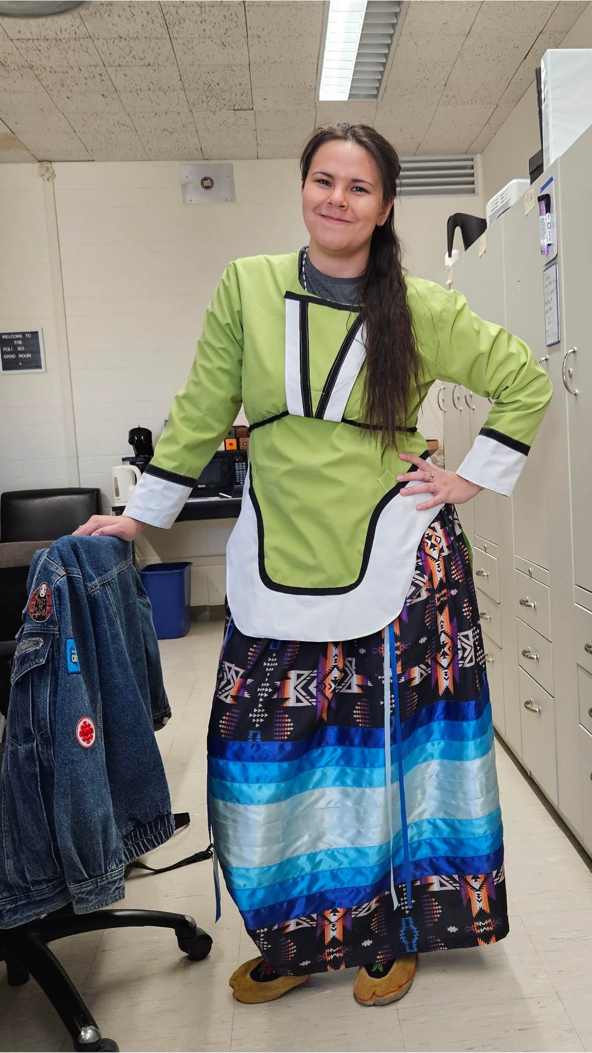 A young Indigenous women standing with her hand on one hip and one hand leaning against a chair. She has long hair worn in a ponytail and she is wearing a black, blue, and white ribbon skirt and moccassins. 