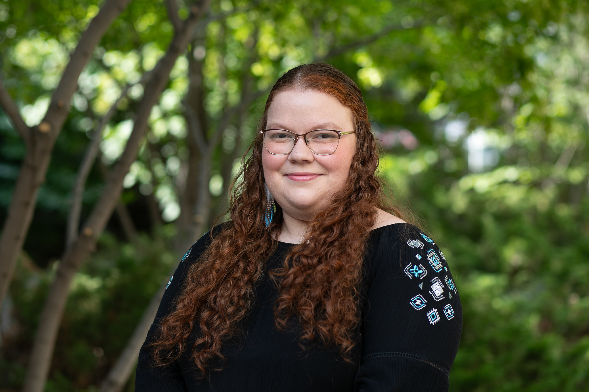 A red-haired woman in black beaded long-sleeve shirt, standing in front of a green, wooded background. 