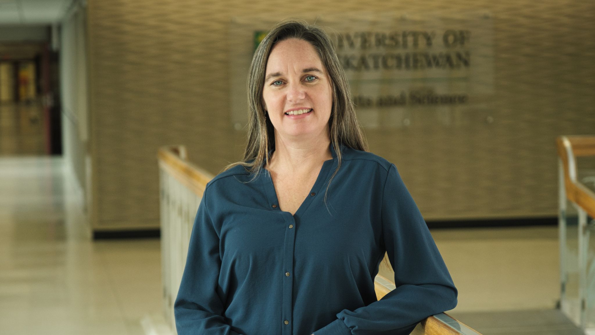 Pictured: Headshot of a caucasian woman with brown hair wearing a blue blouse, standing indoors in front of a University of Saskatchewan sign. 
