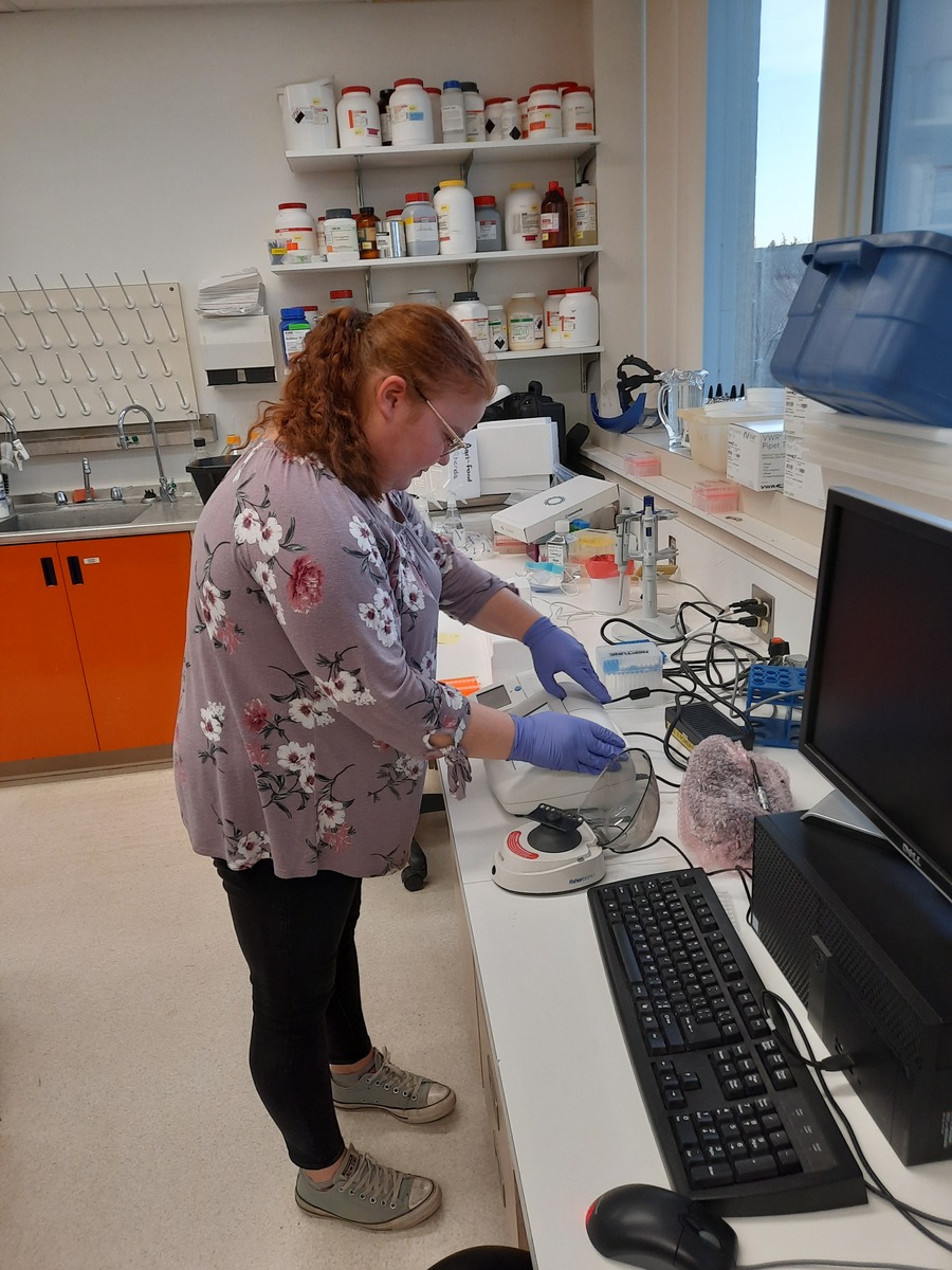 A woman is standing in a lab environment, leaning over a scientific instrument. 