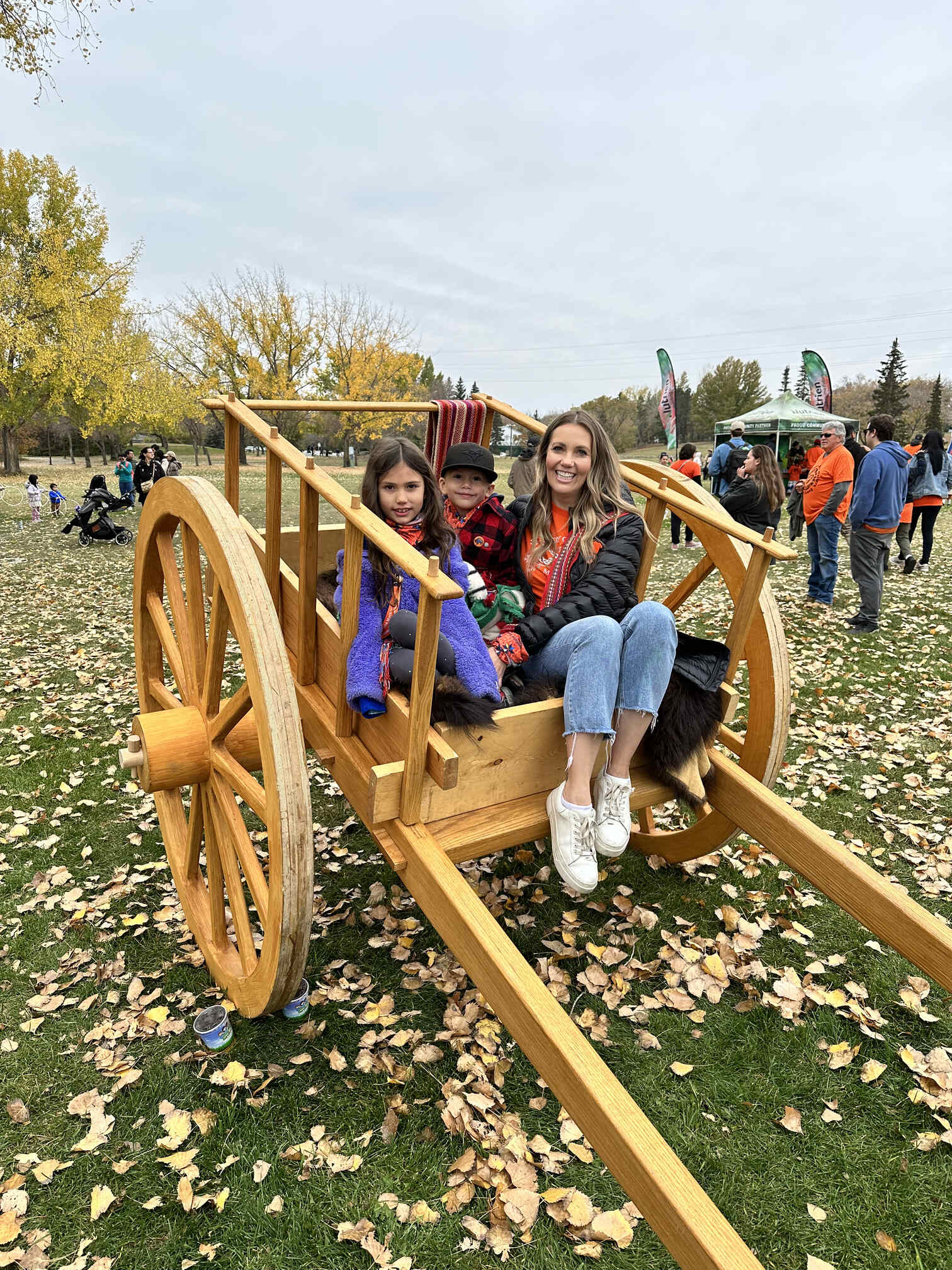 A Métis woman and her two children, a young boy and girl, sitting in a wooden Red River cart in a green field, with autumn leaves on the ground around them.